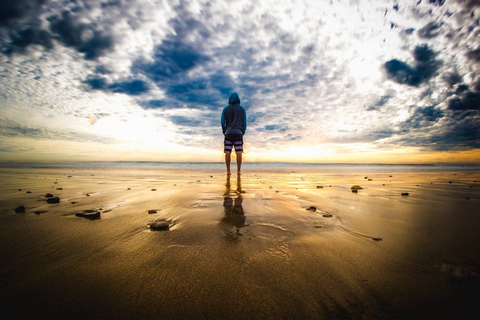 pexels-photo-301952-301952-1 A lone figure stands on a tranquil beach during a vivid sunset, reflecting on the wet sand.